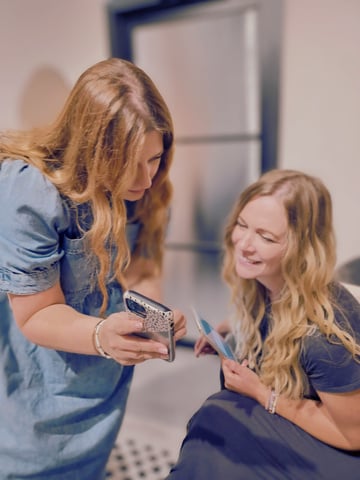 Two white females with long wavy hair looking at a mobile together and smiling.