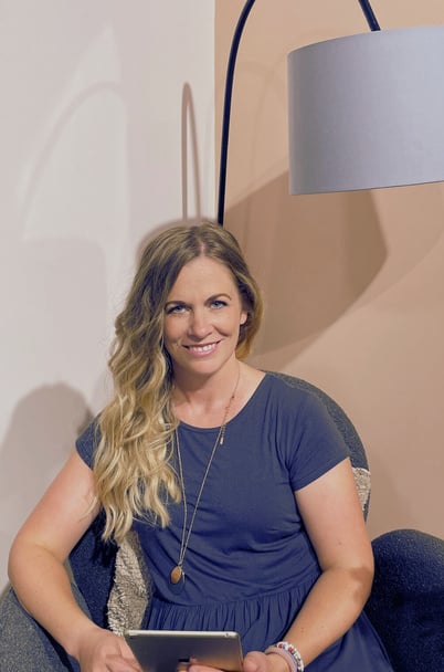 A white female with long wavy blonde hair smiling at the camera and sitting in a chair holding a tablet in front of them.