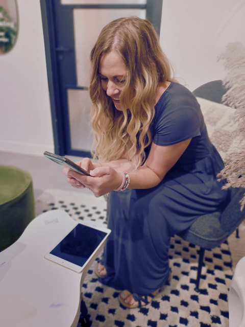 A white female with blond wavy hair sitting on a sofa smiling and looking at her phone.